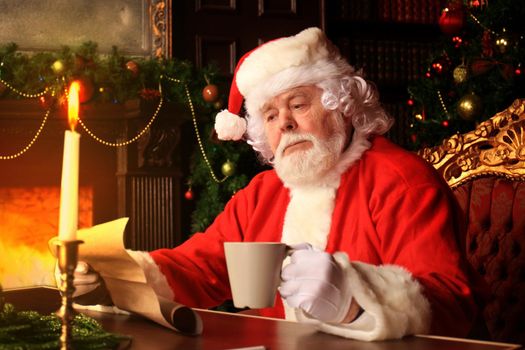 Portrait of happy Santa Claus sitting at his room at home near Christmas tree and reading Christmas letter or wish list.