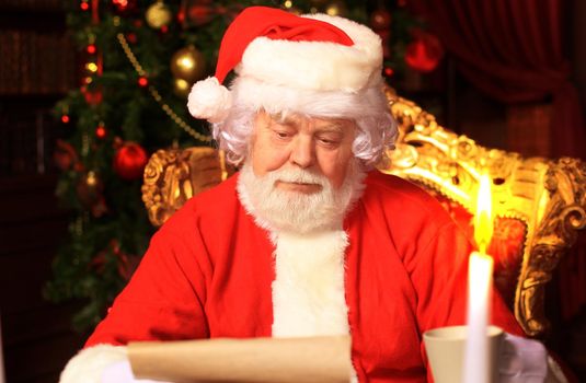 Portrait of happy Santa Claus sitting at his room at home near Christmas tree and reading Christmas letter or wish list.