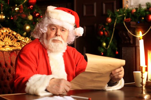 Portrait of happy Santa Claus sitting at his room at home near Christmas tree and reading Christmas letter or wish list.