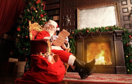 Portrait of happy Santa Claus sitting at his room at home near Christmas tree and reading Christmas letter or wish list.