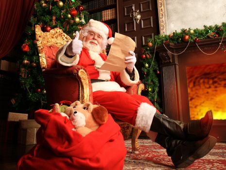 Portrait of happy Santa Claus sitting at his room at home near Christmas tree and reading Christmas letter or wish list.