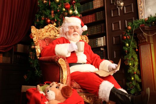 Portrait of happy Santa Claus sitting at his room at home near Christmas tree and reading Christmas letter or wish list.