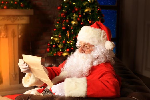 Portrait of happy Santa Claus sitting at his room at home near Christmas tree and reading Christmas letter or wish list.