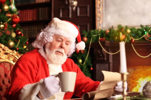 Portrait of happy Santa Claus sitting at his room at home near Christmas tree and reading Christmas letter or wish list.