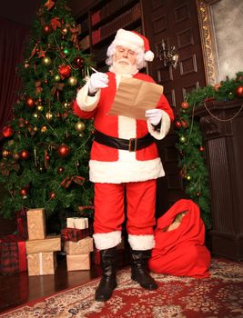 Santa Claus standing at his room at home near Christmas tree and big sack and reading Christmas letter or wish list.