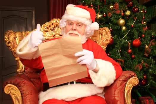 Portrait of happy Santa Claus sitting at his room at home near Christmas tree and reading Christmas letter or wish list.