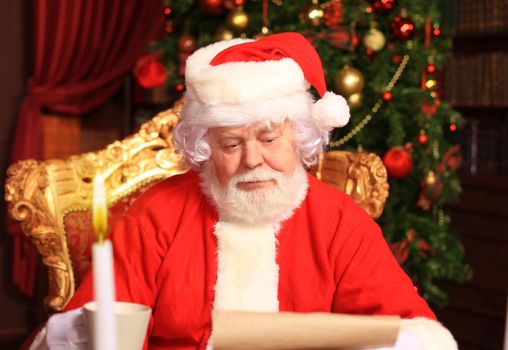 Portrait of happy Santa Claus sitting at his room at home near Christmas tree and reading Christmas letter or wish list.