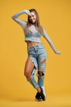 Front view of smiling girl with long brown hair. Woman crossing legs, raising hand and stretching sleeves. Girl wearing short top, ripped jeans on isolated yellow background. Concept of style, beauty