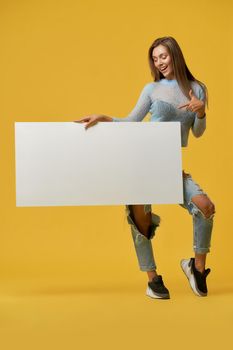 Front view of smiling young stylish brunette girl pointing at white paper sheet in her hand. Fashionable attractive girl with desk posing on yellow studio background. Concept of advertising.