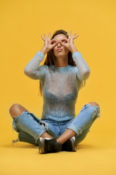 Front view of funny pretty girl sitting with crossed legs, closed eyes, pushed lips showing glasses with fingers on face wearing blue top with long sleeves, ripped jeans.