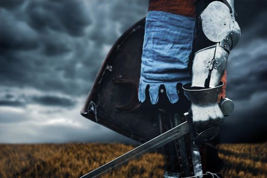 Side close up of incognito medieval knight in armor holding sword and shield. Brave man with cold weapon ready for battle in field, cloudy grey sky, autumn. Concept of warriors.