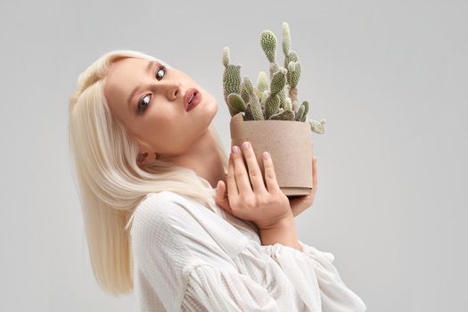 Portrait of beautiful blonde girl with make up and hairstyle wearing white blouse, looking at camera and holding pot with green cactus. Pretty young woman buying plant for house