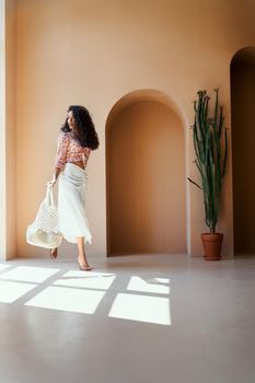 Pretty young female model with long curly hair wearing fashion summer clothes and high heels, holding trendy white purse, looking at big panoramic window. Beautiful girl in a move in studio