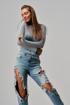 Young pretty girl with long brown hair, closed eyes raising right shoulder, smiling wearing short blue top with long sleeves, ripped jeans on isolated grey background at studio.