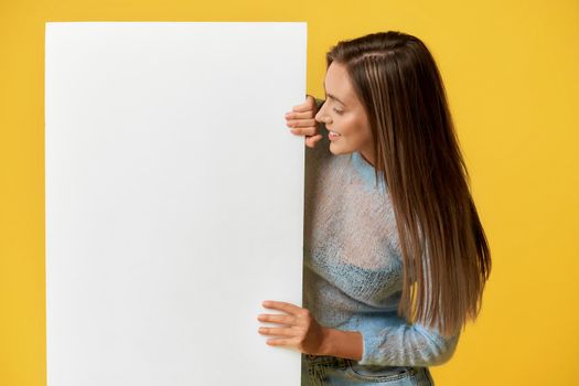 Back view of smiling attractive girl with long brown hair touching, looking at white poster wearing blue top with long sleeves, jeans on isolated yellow background at studio.