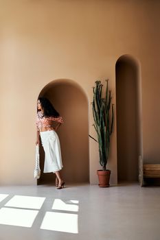 Beautiful girl with curly long dark hair wearing colored crop top, white skirt and high heels standing in decorative arch of the wall near pot of big cactus. Concept of beauty and summer time