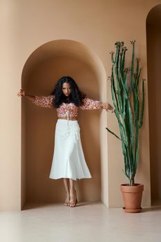 Passionate beautiful young woman with tanned skin in trendy summer clothes standing in niche of wall near big green cactus. Female model with curly hair on high heels posing indoors