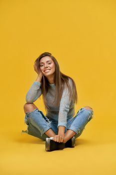 Front view of smiling pretty girl resting cheek in hand sitting with crossed legs looking at camera wearing blue top with long sleeves, ripped jeans.