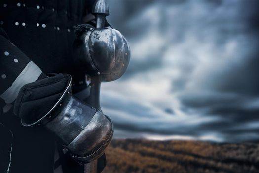 Close up of incognito medieval knight in armor holding sword. Crop of man standing in field and waiting for battle, cloudy weather, overcast, autumn. Concept of warrior.