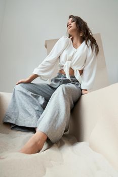 Attractive young lady with wavy long hair in white tied up blouse and grey trousers sitting on the edge of big paper box full of sea sand. Professional model posing on camera.