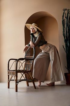 Beautiful blonde girl in big straw hat wearing crop top and wide beige trousers leaning on rattan chair and looking away. Young lady with barefoot standing near wall with decorative niche.