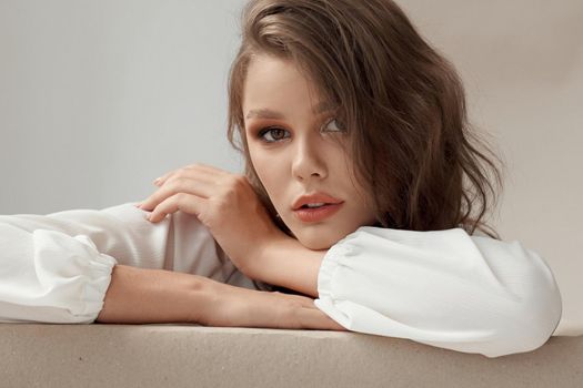 Close up of charming, sensual, young female model with wavy long hair and make up wearing white blouse, looking at camera. Beautiful woman face close up portrait in studio.