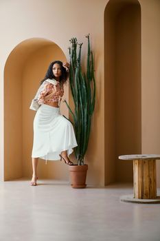 Beautiful brunette girl in colored blouse, white skirt and high heels put her one leg on big ceramic pot with green cactus and lean with one hand on the corner of decorative arch