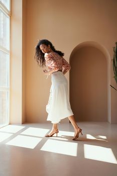 Full length portrait of attractive young model with long curly hair and tanned skin in trendy summer clothes posing on camera near big panoramic window. Concept of beauty and fashion