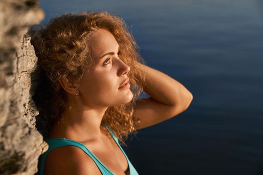 Pretty, attractive girl leaning on rock near water and touching hair. Beautiful model with long curly hair looking up wistfully. Portrait of young charming woman on background of water.