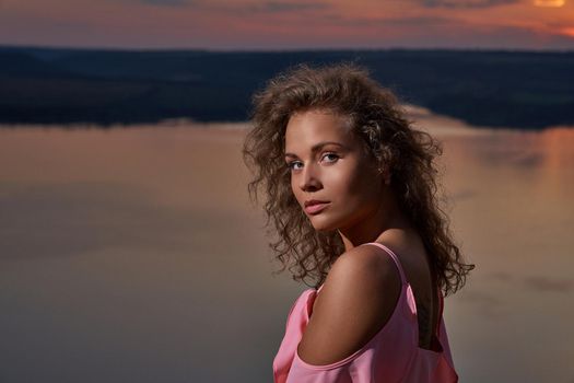 Portrait of seductive girl on background of sundown and water in evening. Sexy, romantic lady wearing in pink dress with long curly hair and bare shoulders looking at camera.