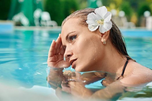 Attractive girl with white flower in hair touching head with hand, looking away. Young caucasian woman relaxing in swimming pool at spa resort. Relaxing concept.