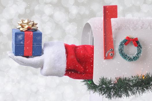 Santa Claus arm holding a small Christmas Present coming out of a mail box with silver bokeh background and snow effect.