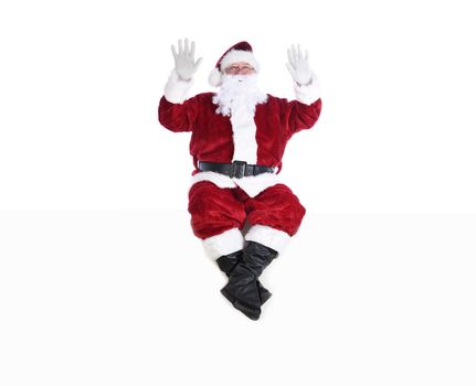 Senior man in traditional Santa Claus Suit sitting on a white wall with both hands in the air.  Isolated on white with copy space.