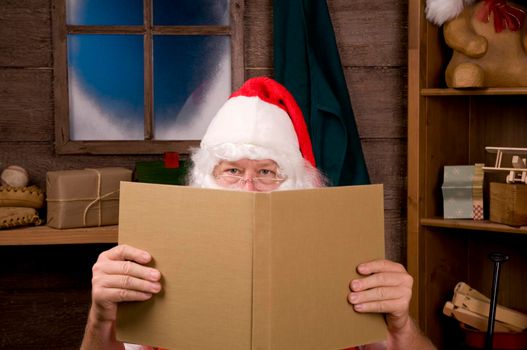 Santa Claus Sitting in His Workshop peering over the top of a large book. Horizontal Composition - Focus on Santa's eyes, slight reflection in window.