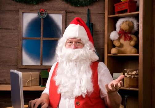 Santa Claus in his workshop with laptop and surrounded by toys and presents.