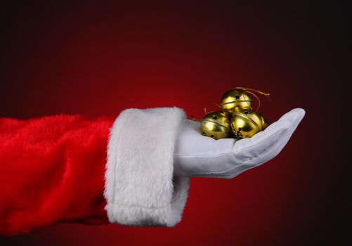 Santa Claus with a handful of gold sleigh bells over a red light ot dark background. Horizontal format showing only hand and arm.