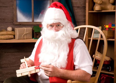 Santa Claus sitting in Rocking Chair in his workshop painting a toy airplane. Horizontal composition.