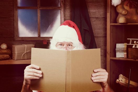 An Instagram style Santa Claus Sitting in His Workshop peering over the top of a large book. Horizontal Composition - Focus on Santa's eyes, slight reflection in window.