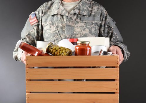 Closeup of a soldier in fatigues holding a wooden box full of canned and packaged food for a holiday charity drive. Horizontal format man is unrecognizable.