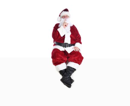 Senior man in traditional Santa Claus Suit sitting on a white wall with a finger to his lips making a Shh Gesture.  Isolated on white with copy space.