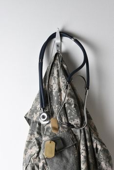 Military Healthcare Concept: A set of dog tags, Stethoscope and camouflage field jacket hanging from a hook on a blank wall.