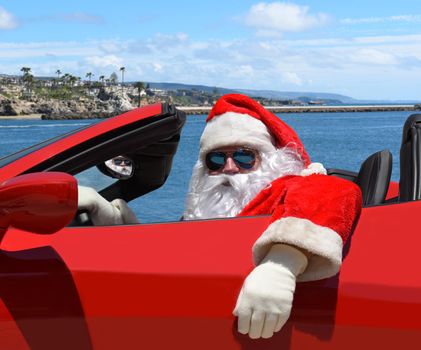 Santa Claus sitting in his red sports car with the ocean and coastline in the background.