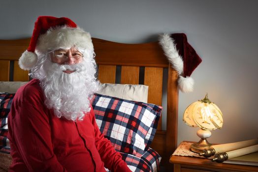 Closeup of Santa Claus wearing a red union suit sitting on the edge of his bed at the North Pole, ready to go to sleep after delivering packages on Christmas eve.