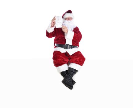 Senior man in traditional Santa Claus Suit sitting on a white wall holding a wrapped Christmas Present.  Isolated on white with copy space.