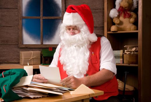 Santa Claus in his workshop reading letters and surrounded by toys and presents.