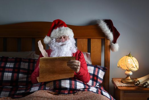 Santa Claus sitting up in his bed working on his naughty and nice list.