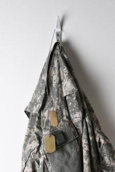 A set of military dog tags and camouflage field jacket hanging from a hook on a blank wall.
