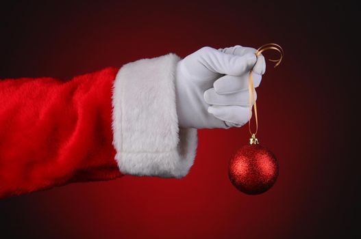 Santa Claus hand holding a sparkly red ornament on a gold ribbon over a light to dark red background. Horizontal format hand and arm only.