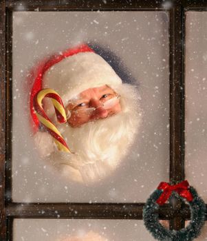 Santa Claus seen through a frosted window holding up a large old fashioned candy cane. It is snowing outside.