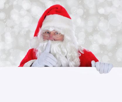 Santa Claus with Blank Sign making shh sign with finger in front of face against a silver bokeh background.